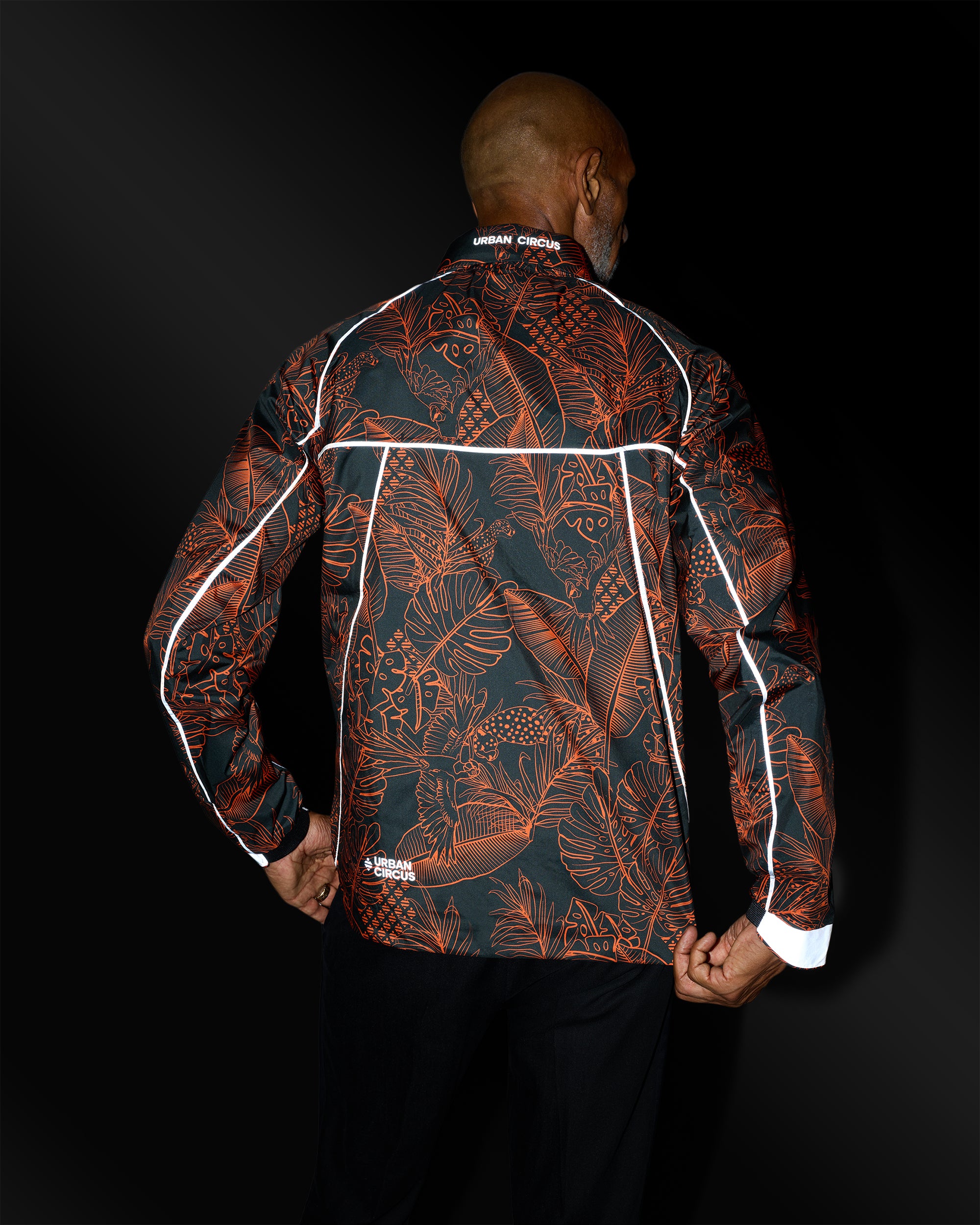 veste légère outdoor respirante noire motifs jungle orange aux détails réfléchissants homme de dos la nuit